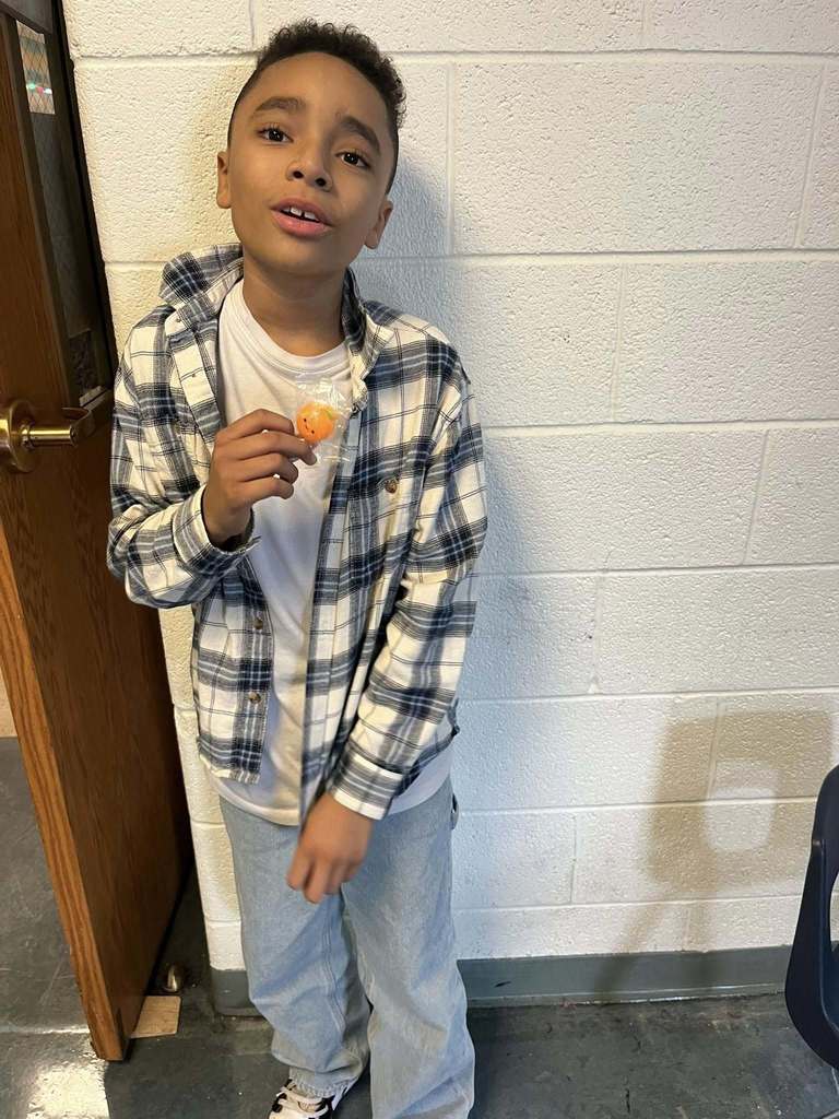 A young boy who is wearing a white shirt and plaid jacket is holding an orange squishy prize he won during bingo.