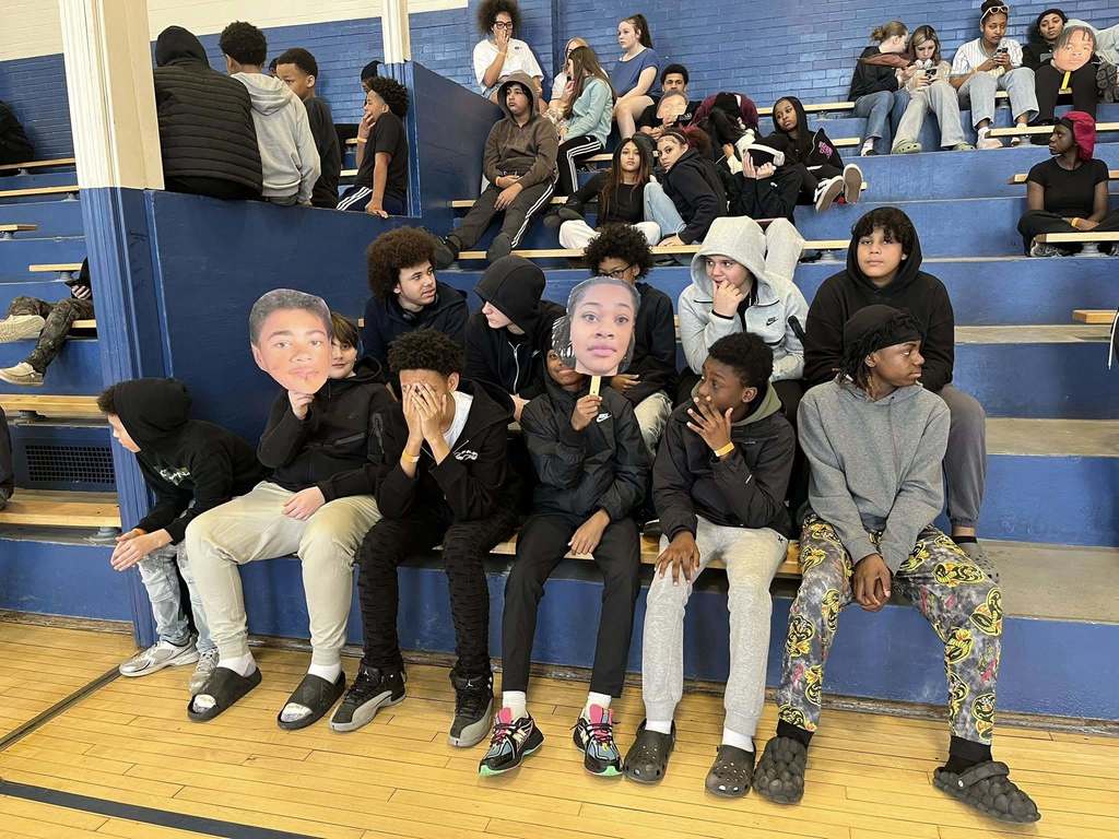 Students sitting on bleachers and two students have cut outs of others who are playing in the basketball game. 