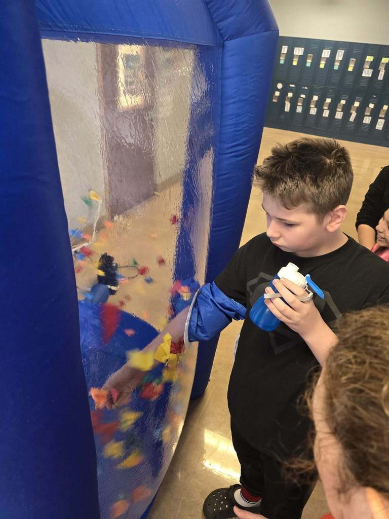 A student has his hand in a blow up tunnel and he is trying to catch as many points as they can which is written on the paper that is flying around in the blow up tunnel. 