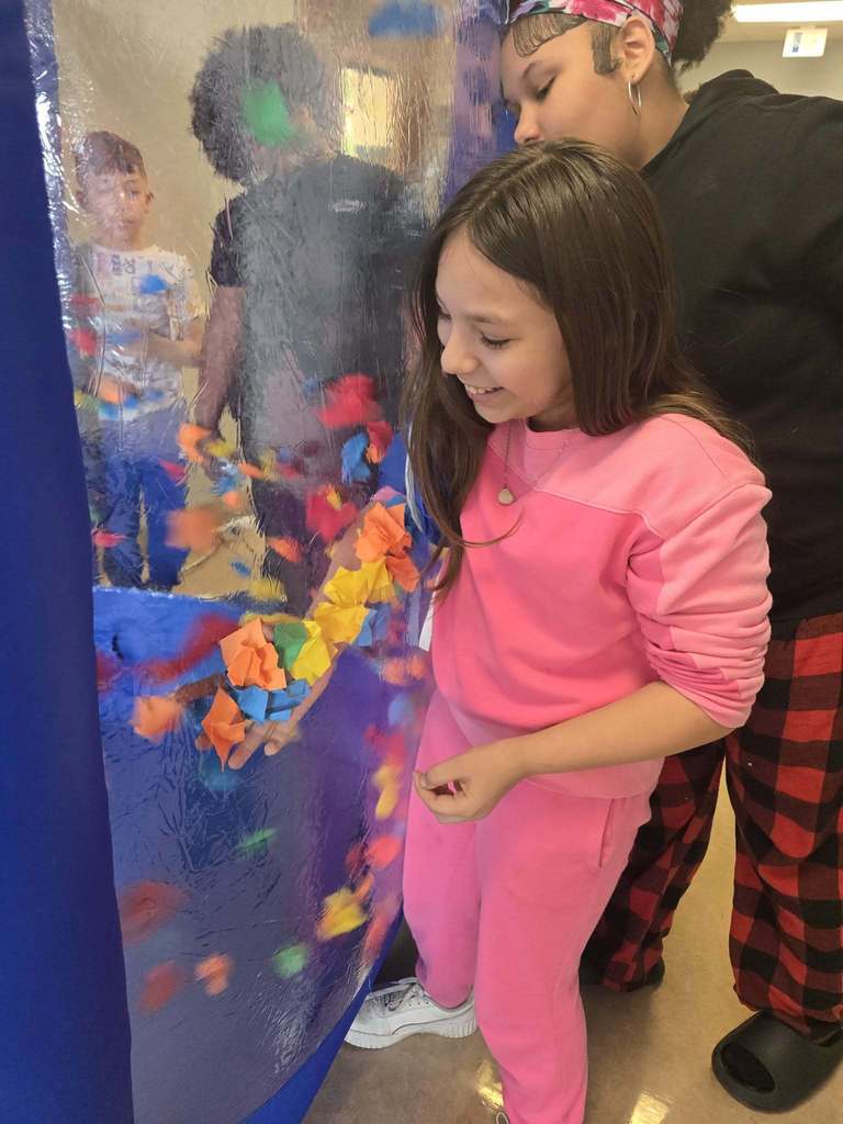 A student has her hand in a blow up tunnel and she is trying to catch as many points as they can which is written on the paper that is flying around in the blow up tunnel. 