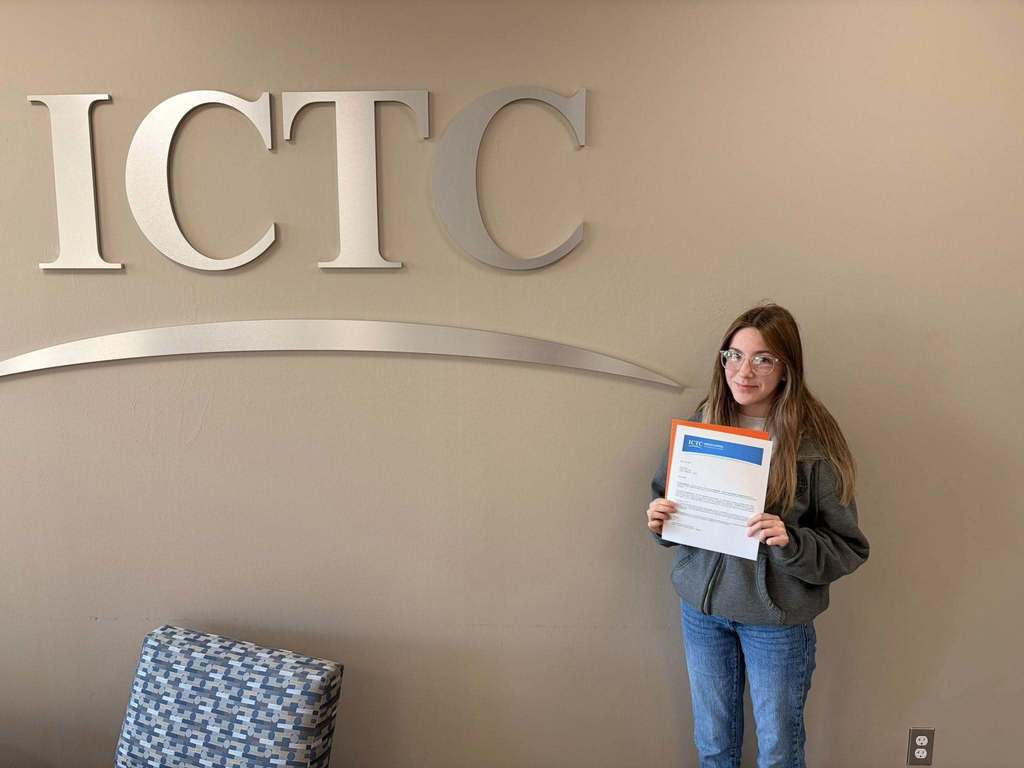 A person in a room stands holding a certificate and smiles in front of a sign reading "ICTC".