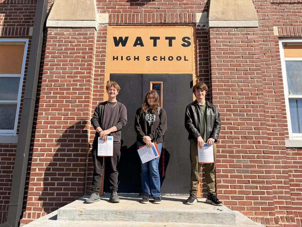 Three students stand in front of Watts High School, holding papers, with a brick wall and school sign in the background.