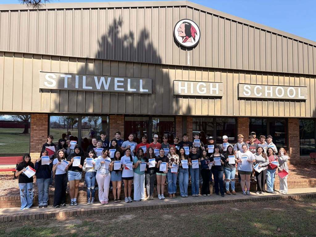 A group of students stands outside Stilwell High School, holding certificates, with a sign reading "STILWELL HIGH SCHOOL" in front of them.