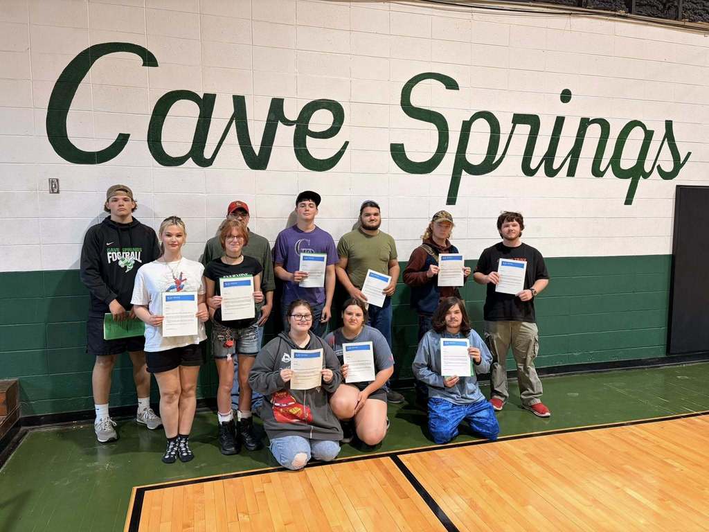 Group of individuals holding certificates pose inside a gym with a "Cave Springs" sign.