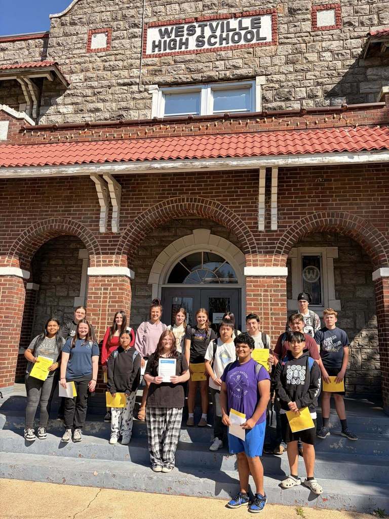 A group of individuals poses in front of Westville High School, holding certificates, with brick walls and arched entrances.