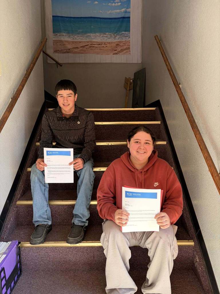 Two individuals sit on a staircase, each holding a certificate, against a backdrop of a beach painting.