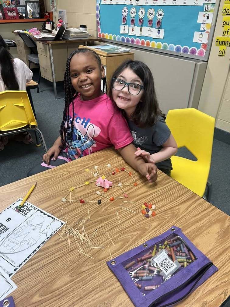 Two young girl leaning on each other in class while they have a project they are working on in front of them on the table. 