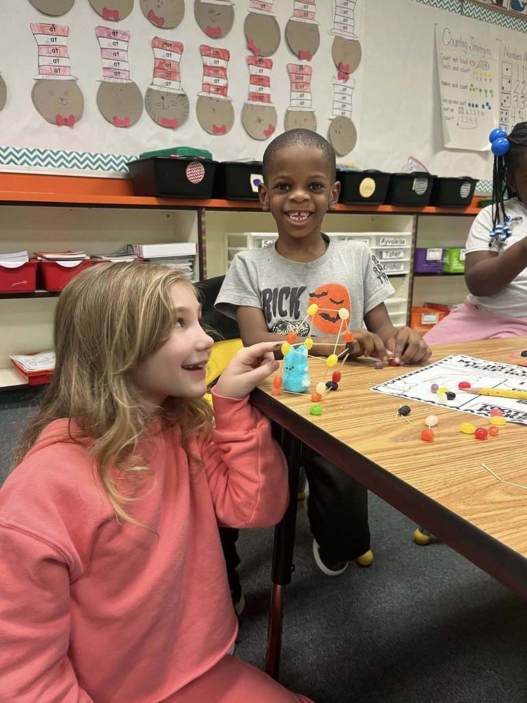 A student pointing at an easter peep inside a house she made of toothpicks and jelly beans while a young boy poses behind the house.