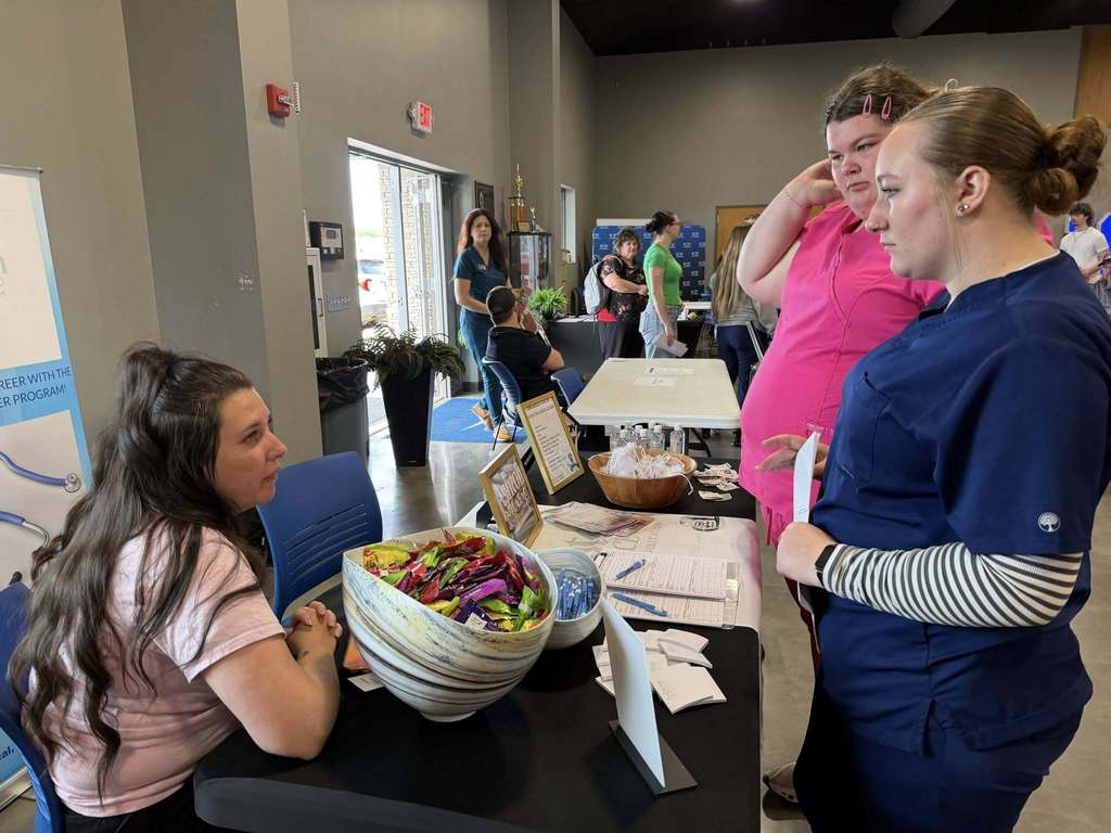 Three people at a table with a bowl of fruit; one person seated, another standing, and a third speaking.
