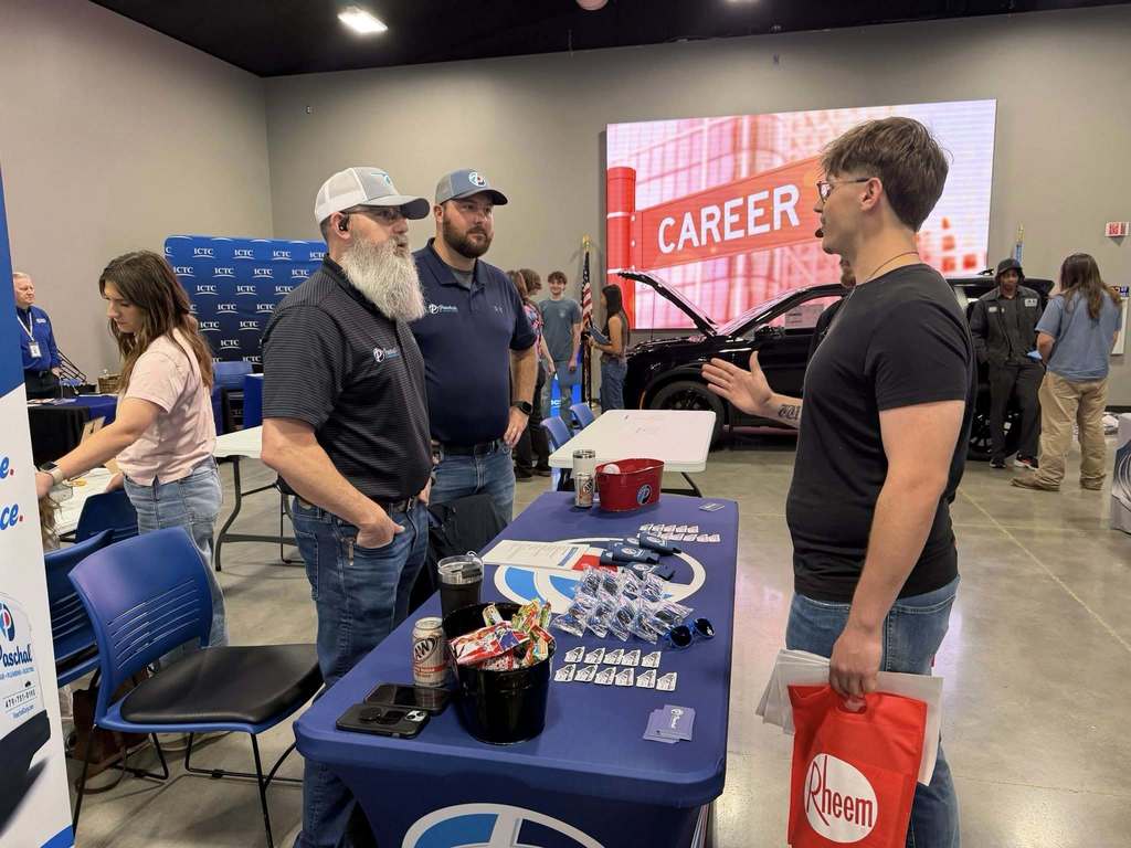 People gather at a table displaying items and a red bag. A man with a beard and a hat converses with another man.