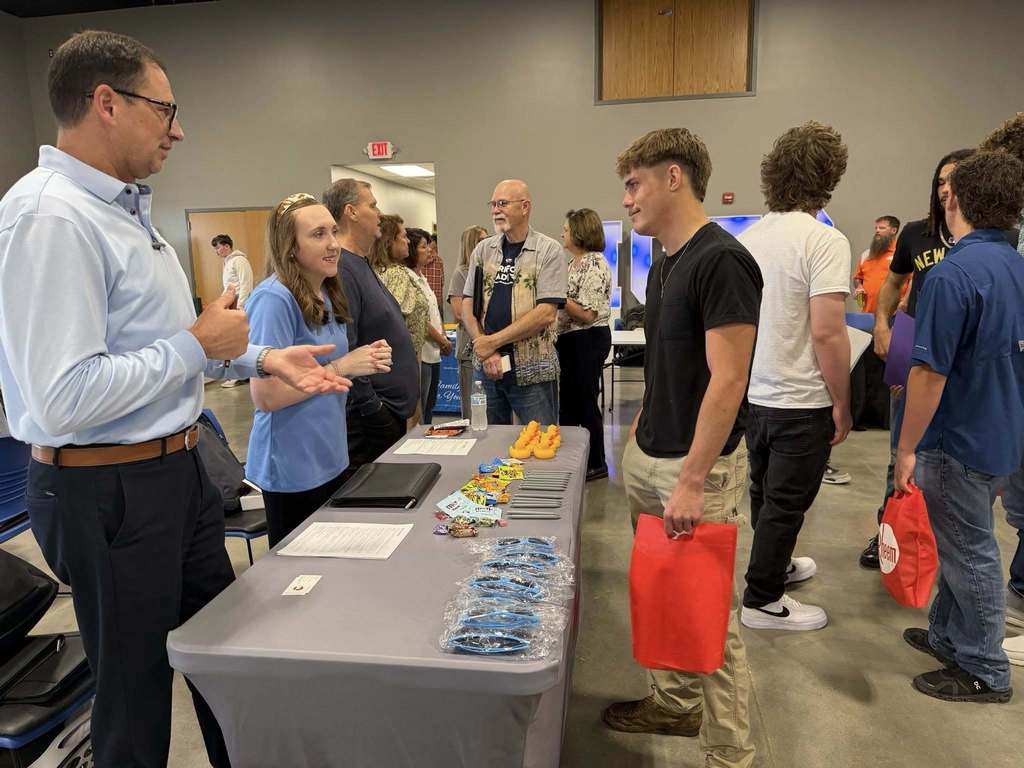 Several people engage in conversation at a table with items on it, in a room with white walls.