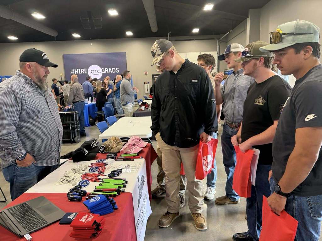 People examine items on a table with a red tablecloth, some holding red bags. They stand in a room with white walls and overhead lighting.