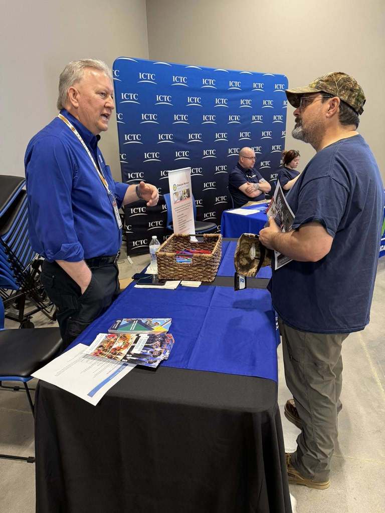 Two men converse at a table with blue cloth. One wears a blue shirt and cap, the other in blue with a lanyard. Behind them, a blue backdrop displays text.