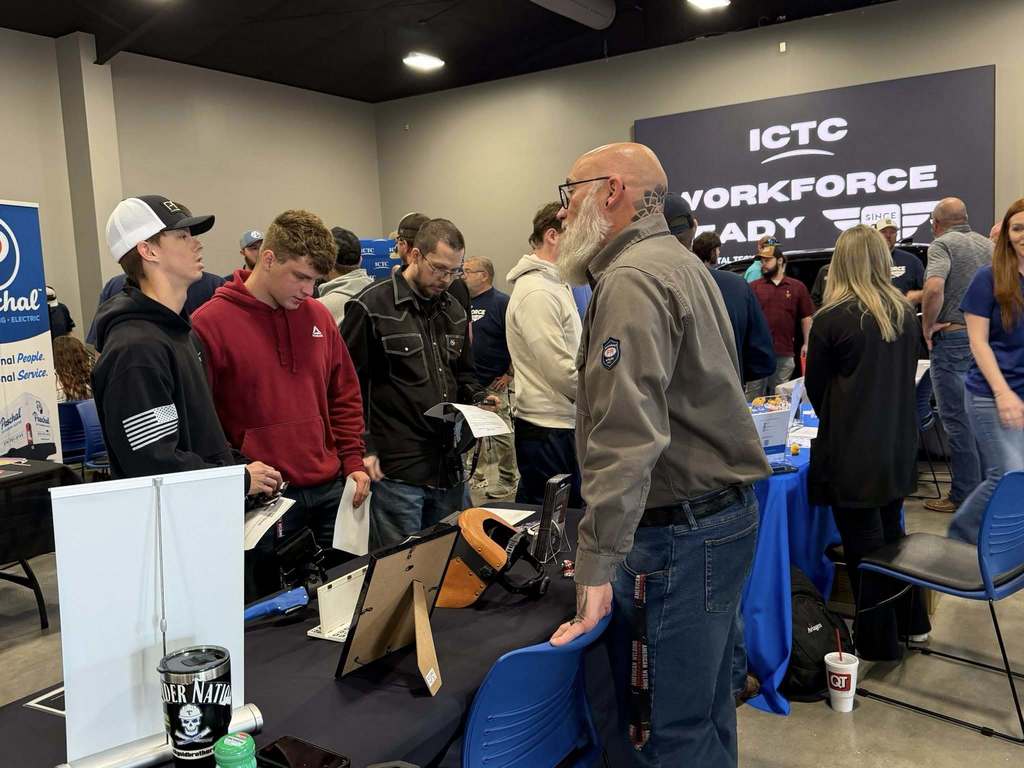Indoor event with tables and chairs; people gather around a booth with a banner saying "ICTC YORKFORCE FACY".