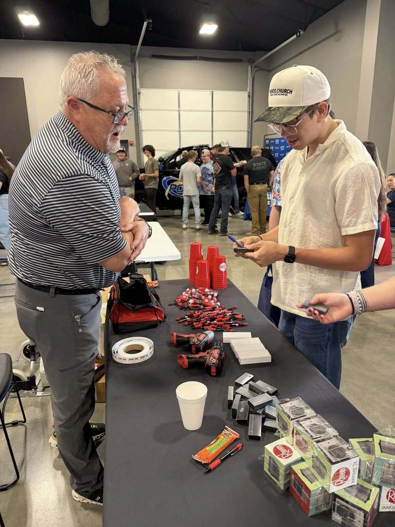 Two men at a table with various items. One wearing glasses, the other with a cap. Multiple people walk behind.