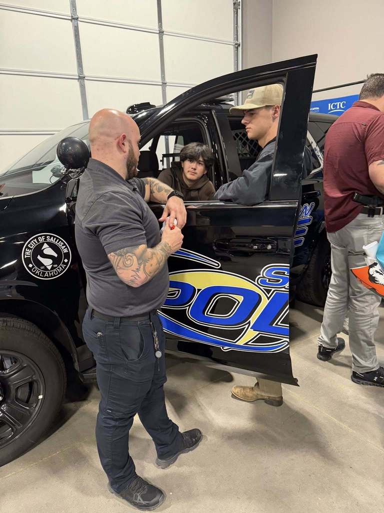 Three men, one with tattoos, standing by a police vehicle with open door. One sits inside. Background is a garage.