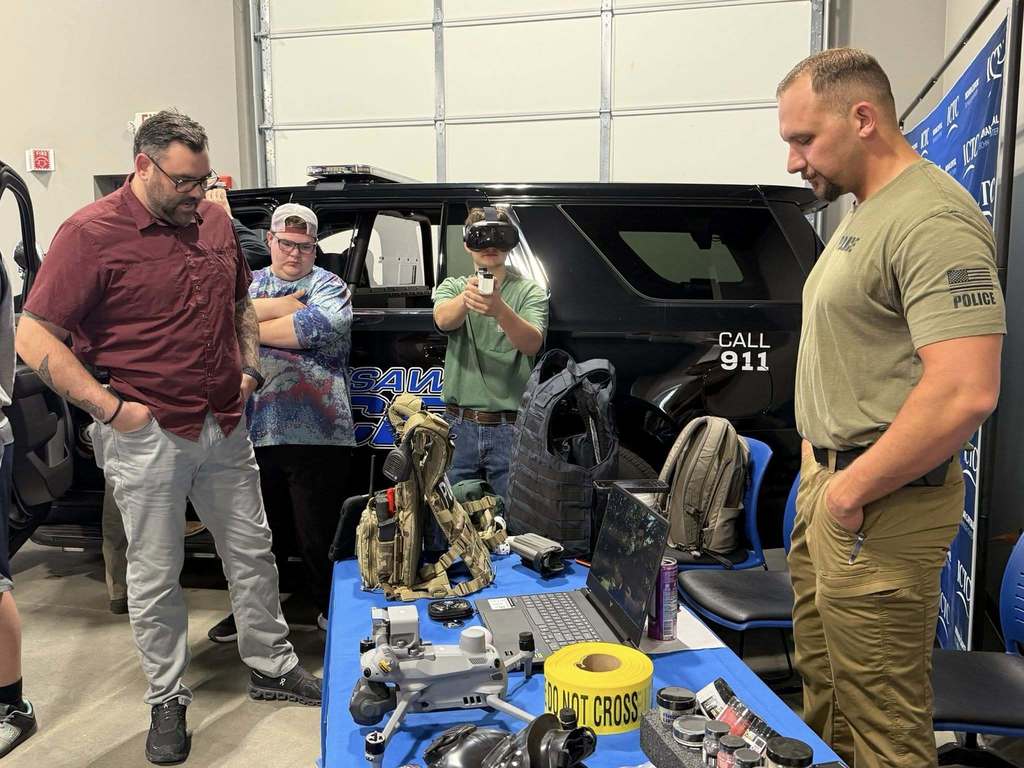 Three men, one in a red shirt and two in green, stand near a black SUV. A table holds various items, including a laptop.