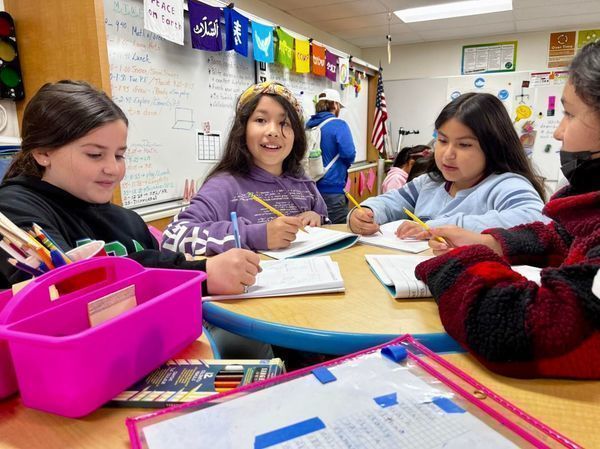 students in classroom at table smiling and writing papers