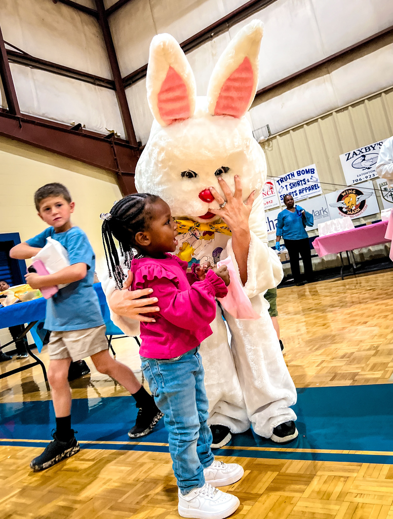 A young girl in pink holds a bunny mascot, while a boy in blue holds a book nearby. They are in a gym.