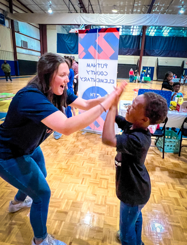 A woman in blue exercises with a child in black inside an indoor court. A banner is behind them.