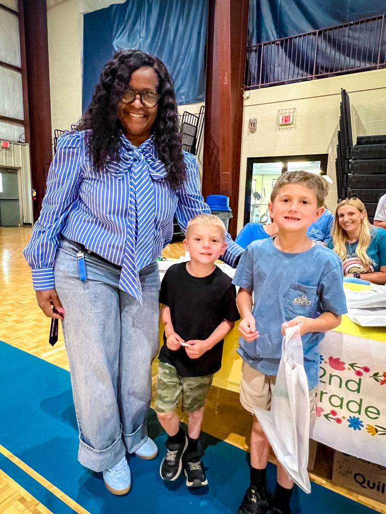 A woman in a blue striped shirt and jeans stands next to two young boys. They are all smiling and standing on a blue carpet.