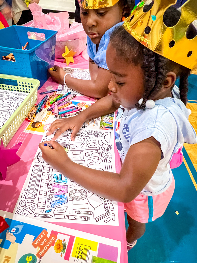 Two young children coloring at a table. One child wears a crown, and both have crayons and coloring books.