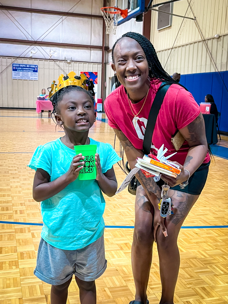 A woman and a child stand in a gym, smiling. The child holds a green cup and a book.