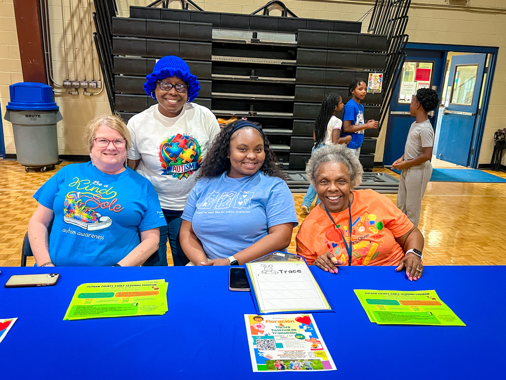 Four adults, smiling, seated at a table with materials. Behind them, a gym with a blue door.