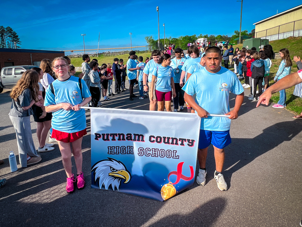 People in blue shirts and shorts, including two holding a banner, walk in a parade. Behind them, a crowd is visible.