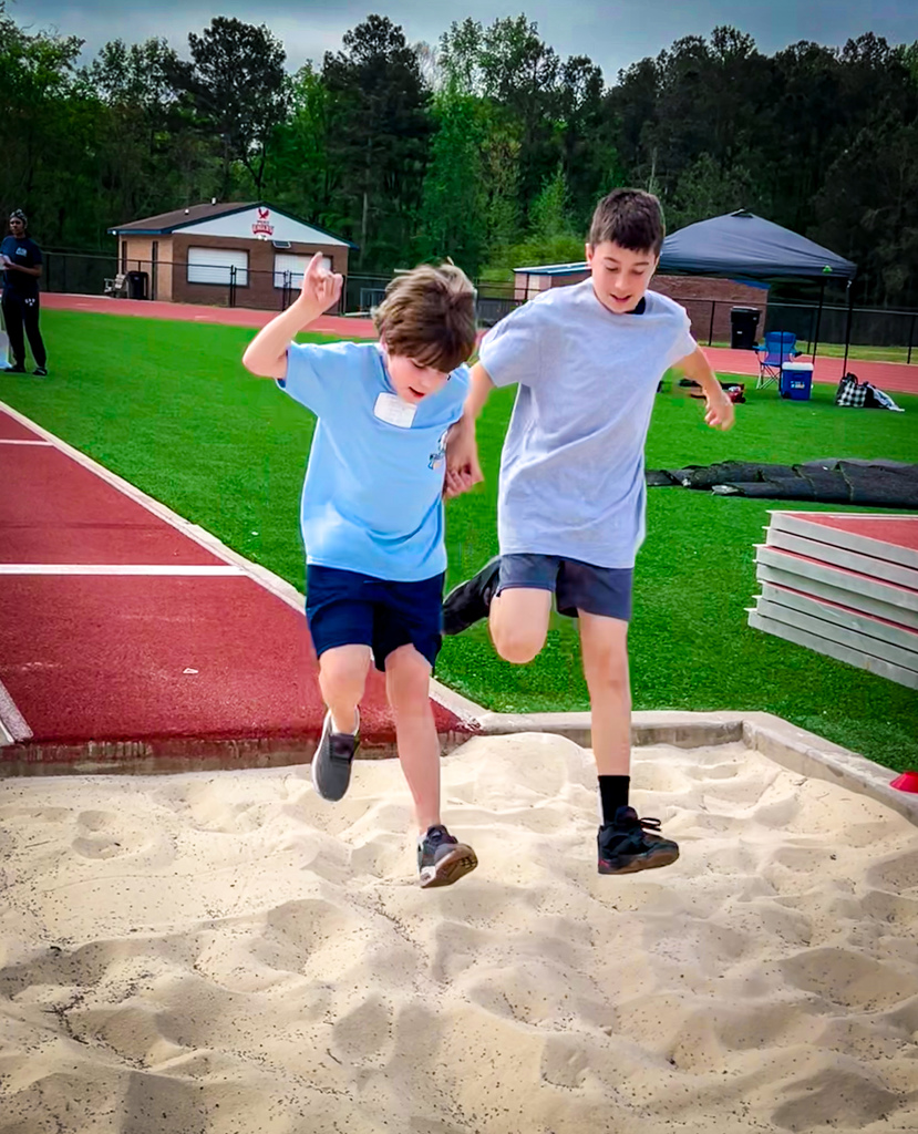 Two children, one jumping, the other assisting, on a track with a sand pit, a red track, and green grass.