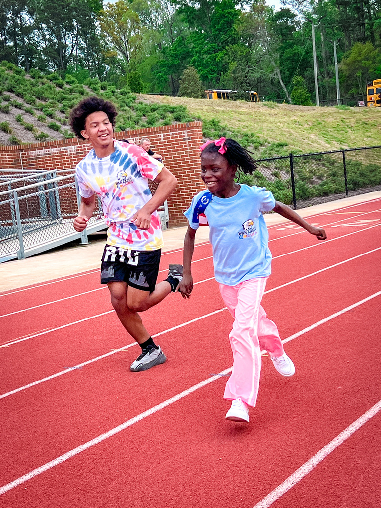 Two children run on a track, one in colorful attire, the other in blue, on a sunny day.