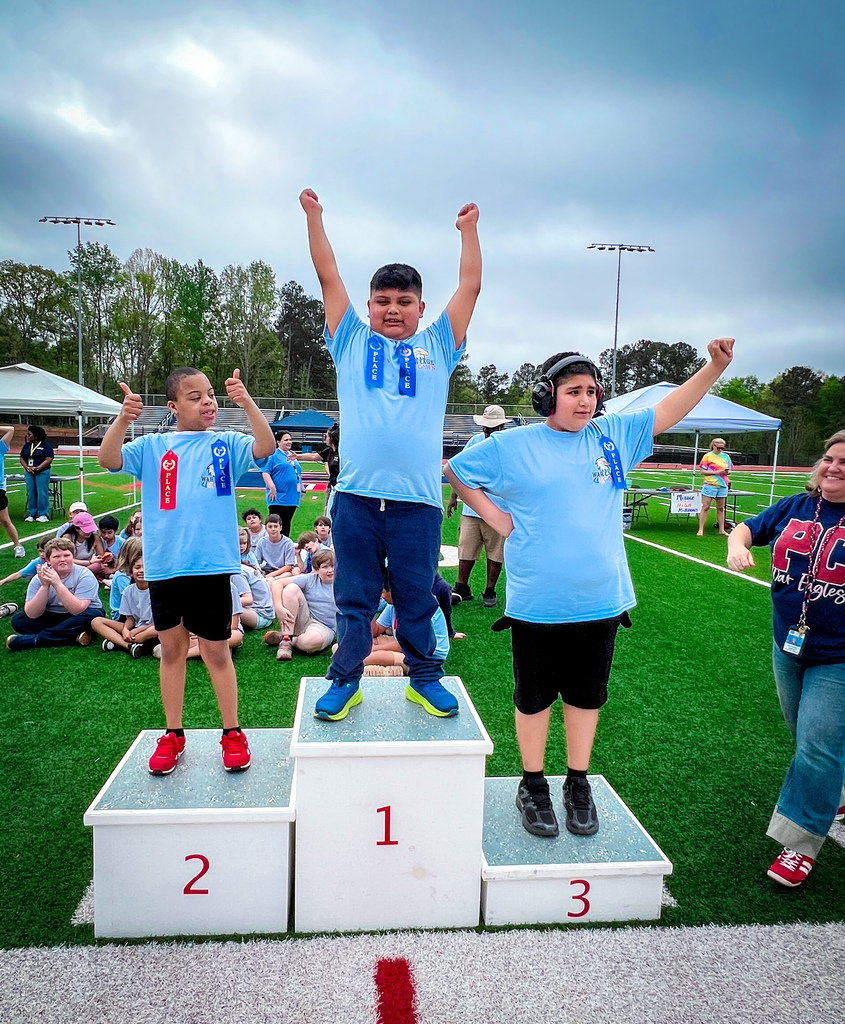 Three young people stand on a podium with 1, 2, and 3 markings. They raise their hands, and there are spectators in the background.