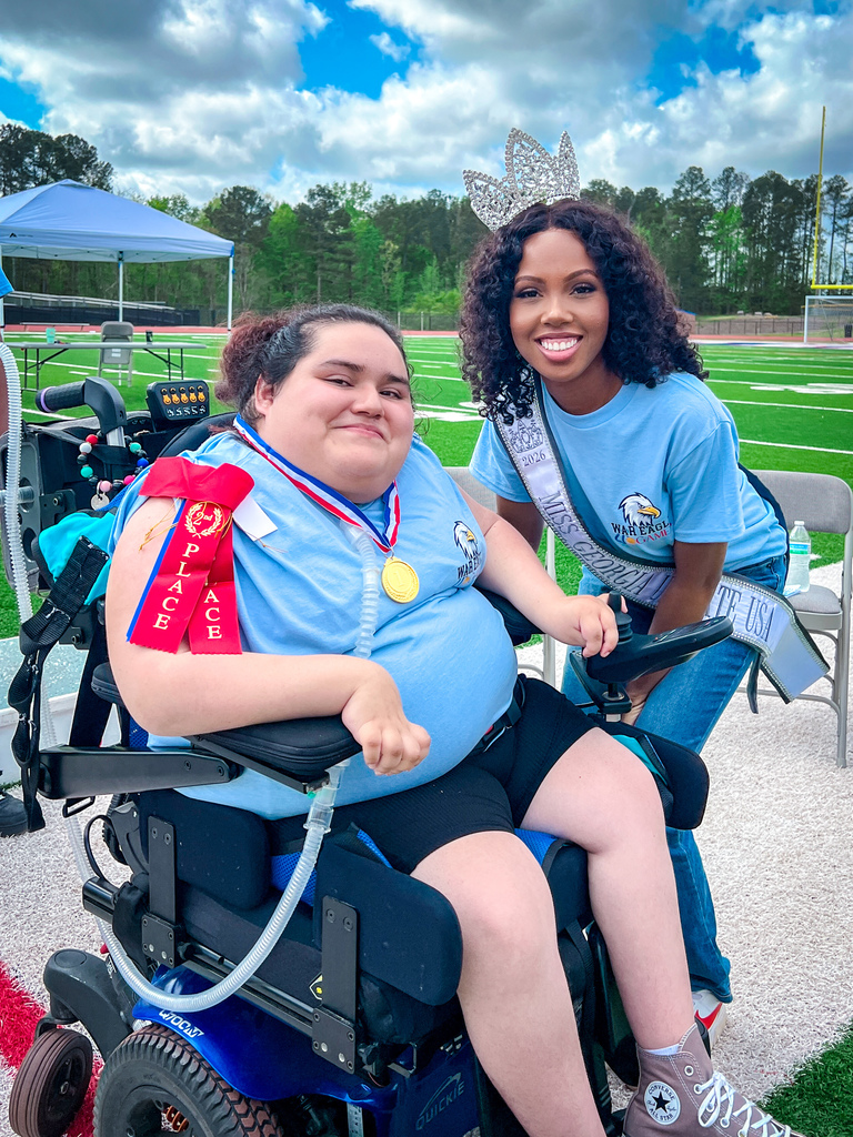 Two women wearing blue shirts, one with a sash and medal, seated in a wheelchair on a field.