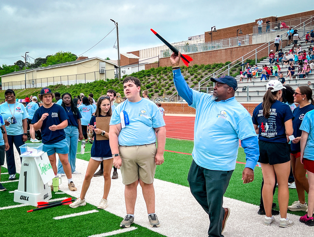 Stadium with audience in stands, a man holds a red rocket. People in blue shirts and shorts, some wearing hats.