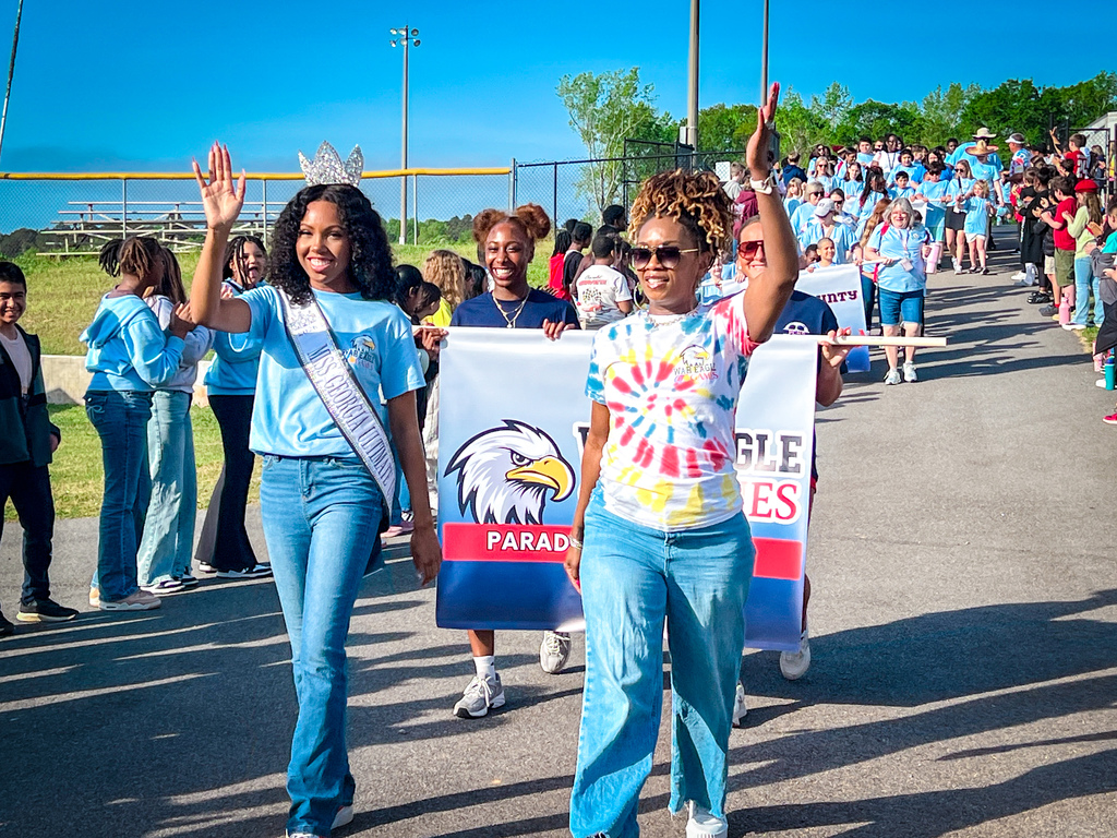 Two women in light blue jeans and white sneakers wave to a crowd on a sunny day.