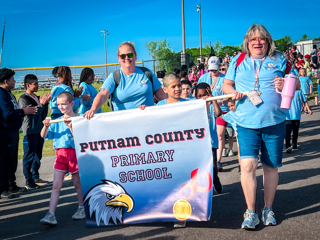 Two adults lead a banner with "Putnam County Primary School" text, an eagle image, and a sun. Children walk behind them, some with hats, in an outdoor setting.