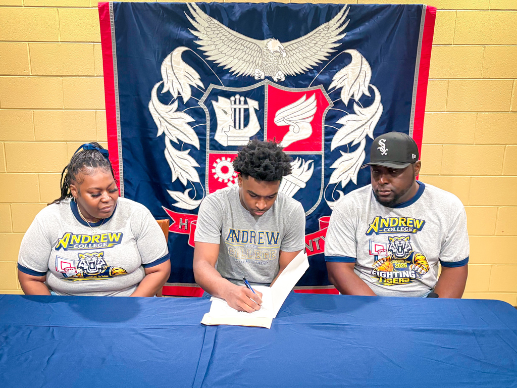 Three people sit at a table, one signing a paper with a pen. Two women and one man wear Andrew shirts. Behind them, a banner hangs.