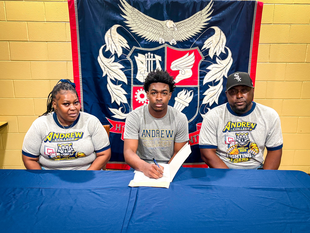 Three people sit at a table with a blue cloth. Two are wearing gray shirts and one wears a black cap. They are signing papers.