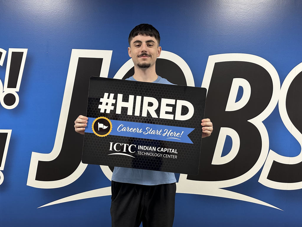 Person stands in front of a blue and white sign, holding a black sign that reads "#HIRED".