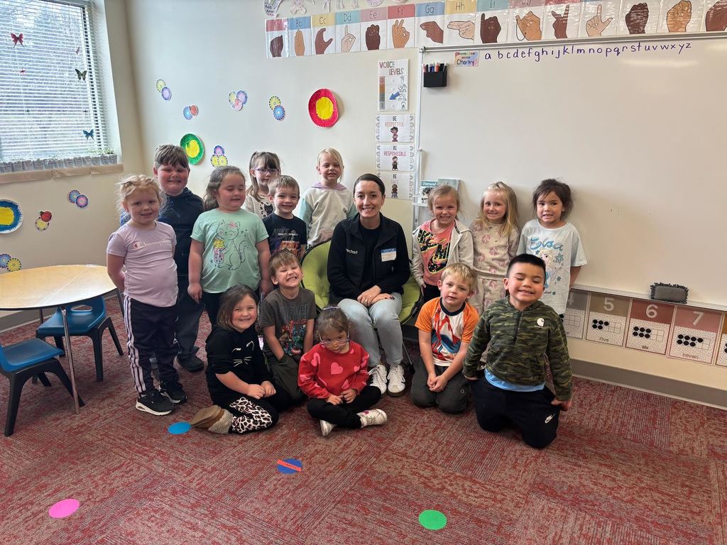 A group of approximately 13 young children and one adult sit and stand together in a brightly lit classroom. The adult, identified as Darcie Brandt, sits in the center smiling, with the children gathered around her—some standing behind and others sitting or kneeling on the carpet in front. The children appear to be preschool age and wear casual, colorful clothing. Most are smiling and facing the camera.  The classroom includes a red carpet, a whiteboard displaying the alphabet, and posters showing hand signs for each letter. Colorful circular artwork decorates the walls, and a window with blinds lets in natural light. A small table and chairs are visible to the left.