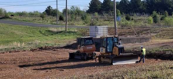 A construction site with a bulldozer parked in the dirt. A worker in a safety vest is standing nearby.