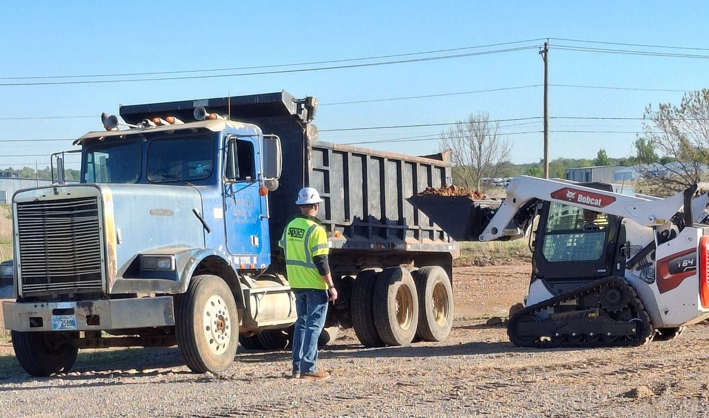 A person in a reflective vest stands near a blue dump truck and a white backhoe on a dirt surface.