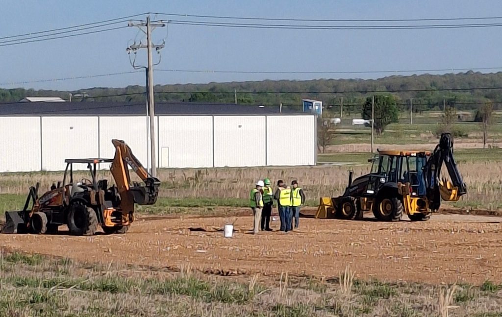 A few people in yellow vests are standing near three large machinery vehicles on a dirt field. Behind them is a large white building.