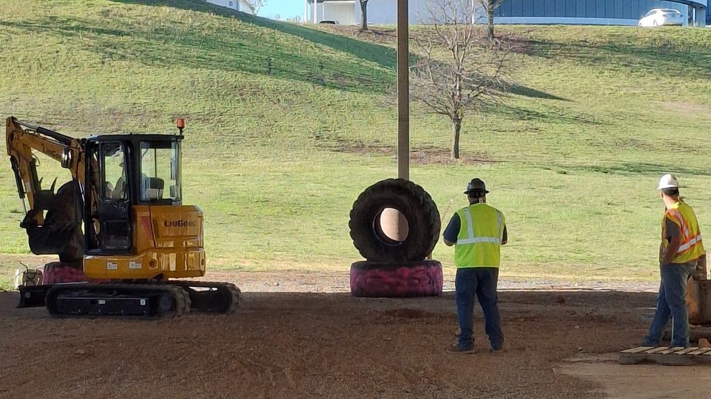 Two workers in yellow vests and white hats stand on a dirt field near a yellow excavator. A large tire sits on a red platform.