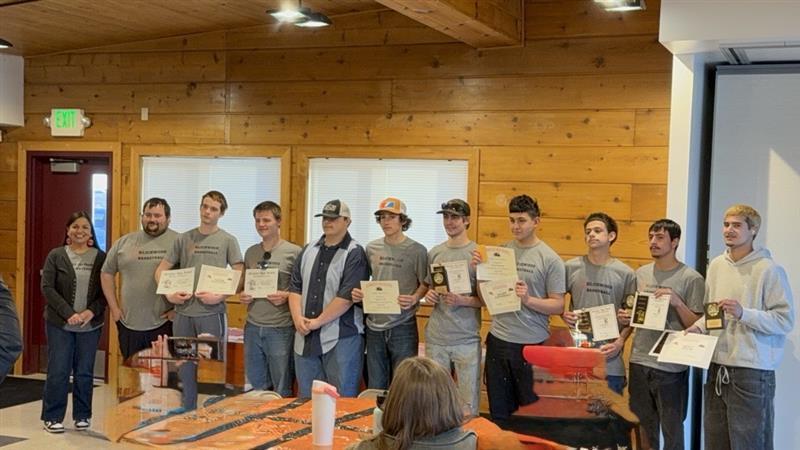 Basketball players standing in a line, with the coach(es) at the left end. All players are holding their awards up.