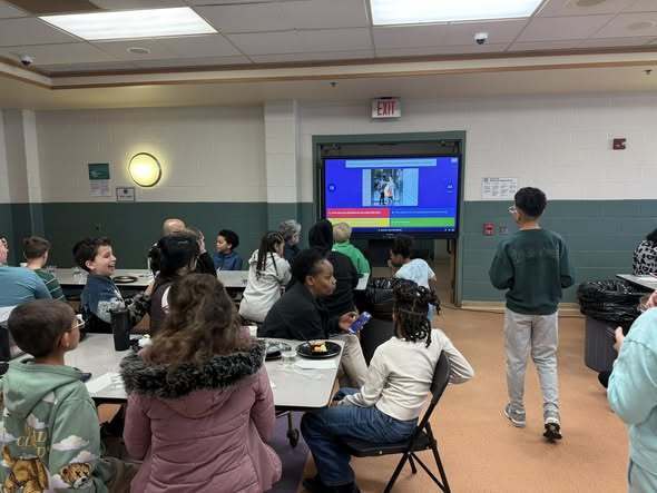 People sitting in a cafeteria and looking at a tv screen. 