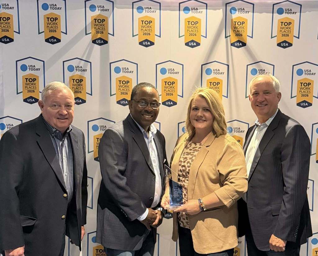 Four adults stand in front of a wall with logos. A woman in the center holds a trophy.