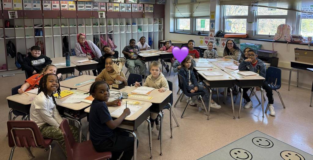 Students and teacher posing in a classroom at their desks while one student holds a trophy.  
