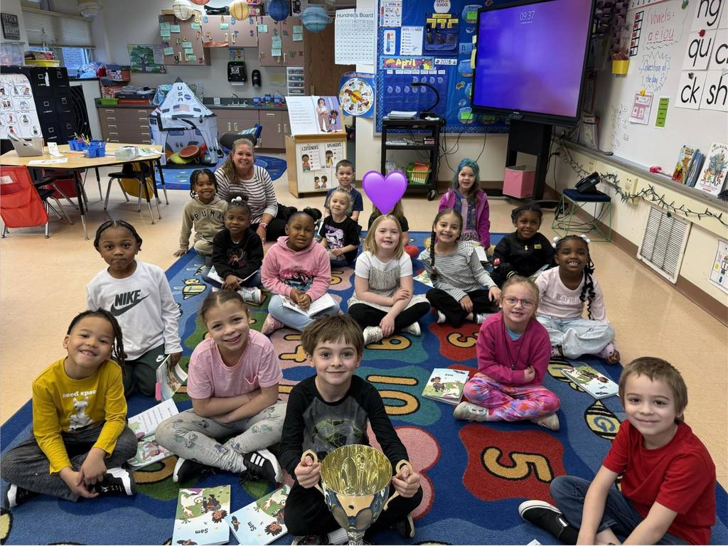 Students and teacher posing in a classroom while one student holds a trophy. 