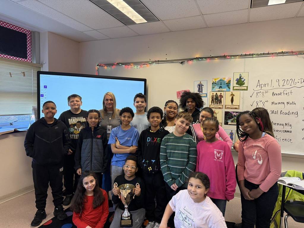 Students and teacher posing in a classroom while one student holds a trophy. 